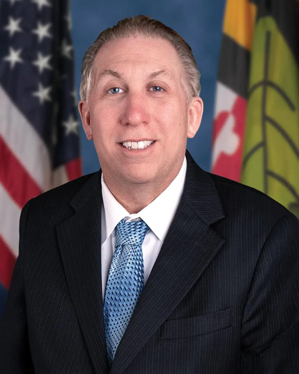 Mike Hart in a dark suit and blue tie, standing in front of the American, Maryland, and Calvert County flags.